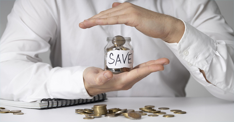 A person is holding a jar filled with coins and a few more pennies on the table, along with a notebook. 